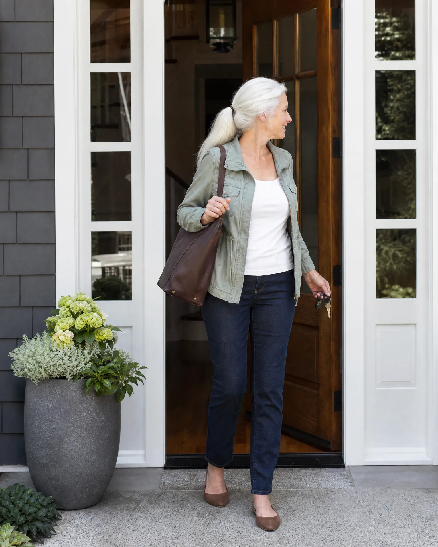 A woman with white hair in a ponytail, wearing a green jacket, white top, dark jeans and brown flats, stands in an open wooden doorway holding keys and a brown shoulder bag, beside a gray planter with flowering plants.