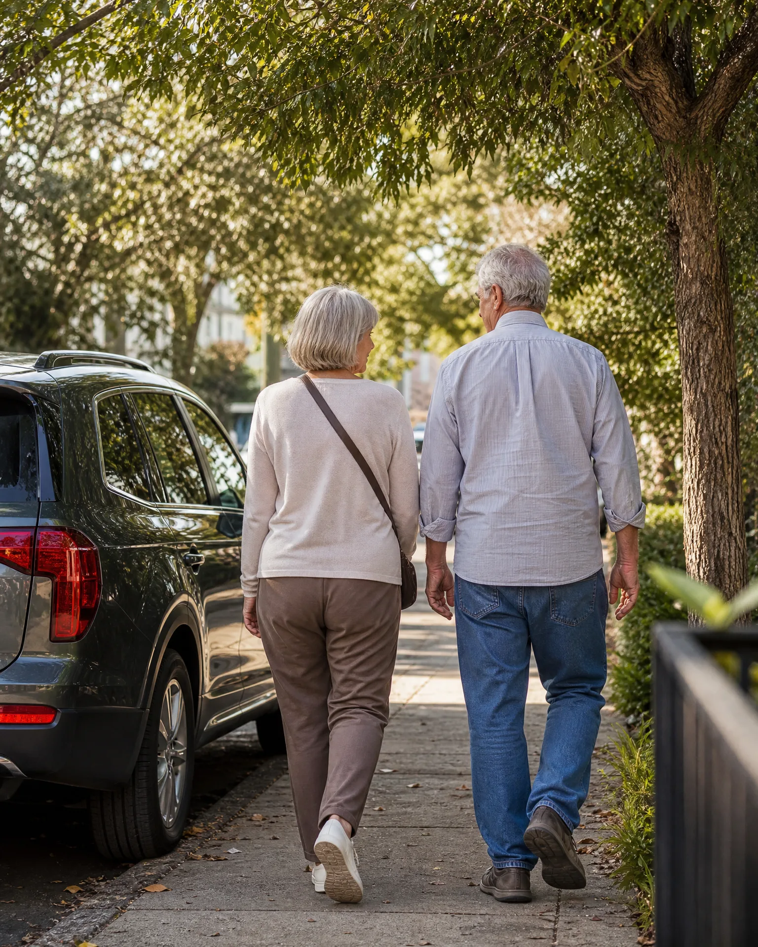 Two gray-haired people walk away from the camera along a tree-shaded concrete sidewalk, with a dark parked SUV at the curb on the left and hedges on the right.