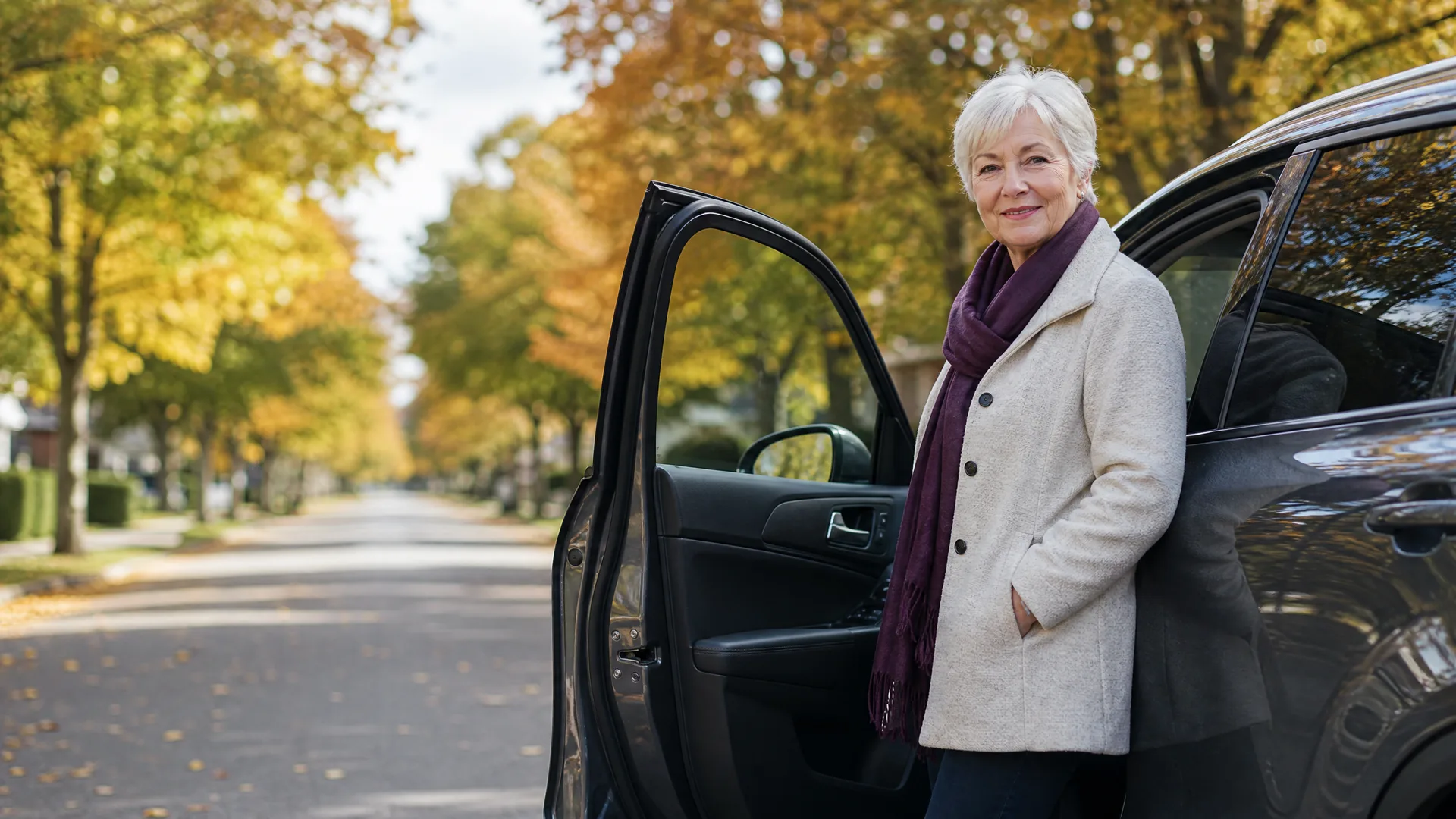 A woman with short white hair in a cream coat and dark purple scarf stands beside the open door of a dark SUV on a tree-lined residential street with autumn foliage.