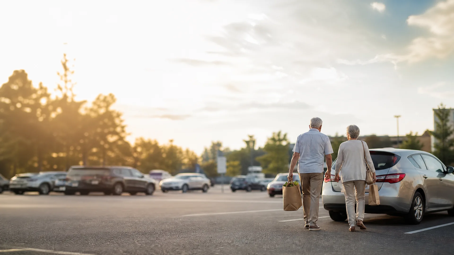 An older man and woman seen from behind walk across a parking lot toward a silver car, the man carrying a paper grocery bag, with parked cars and trees under a bright hazy sky behind them.