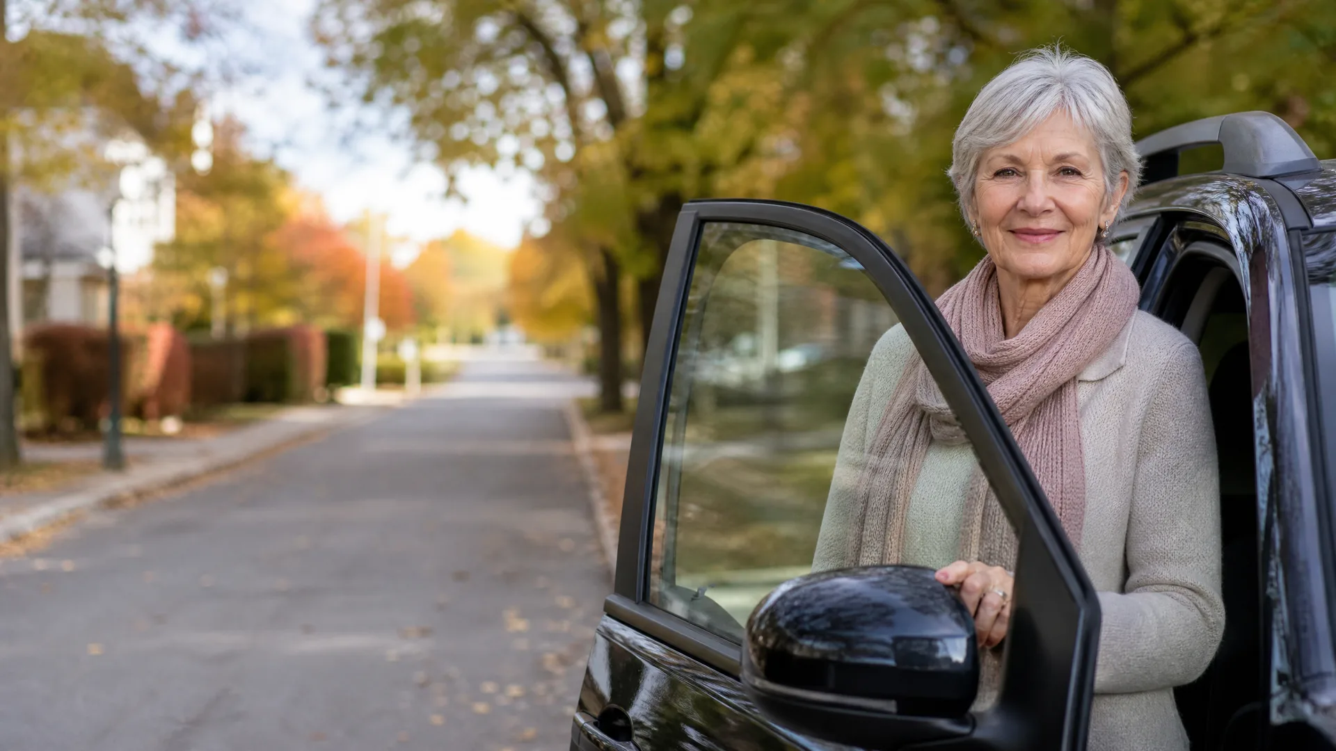 An older woman with short gray hair, wearing a beige cardigan and pink scarf, stands beside the open door of a dark car on a residential street with autumn trees.