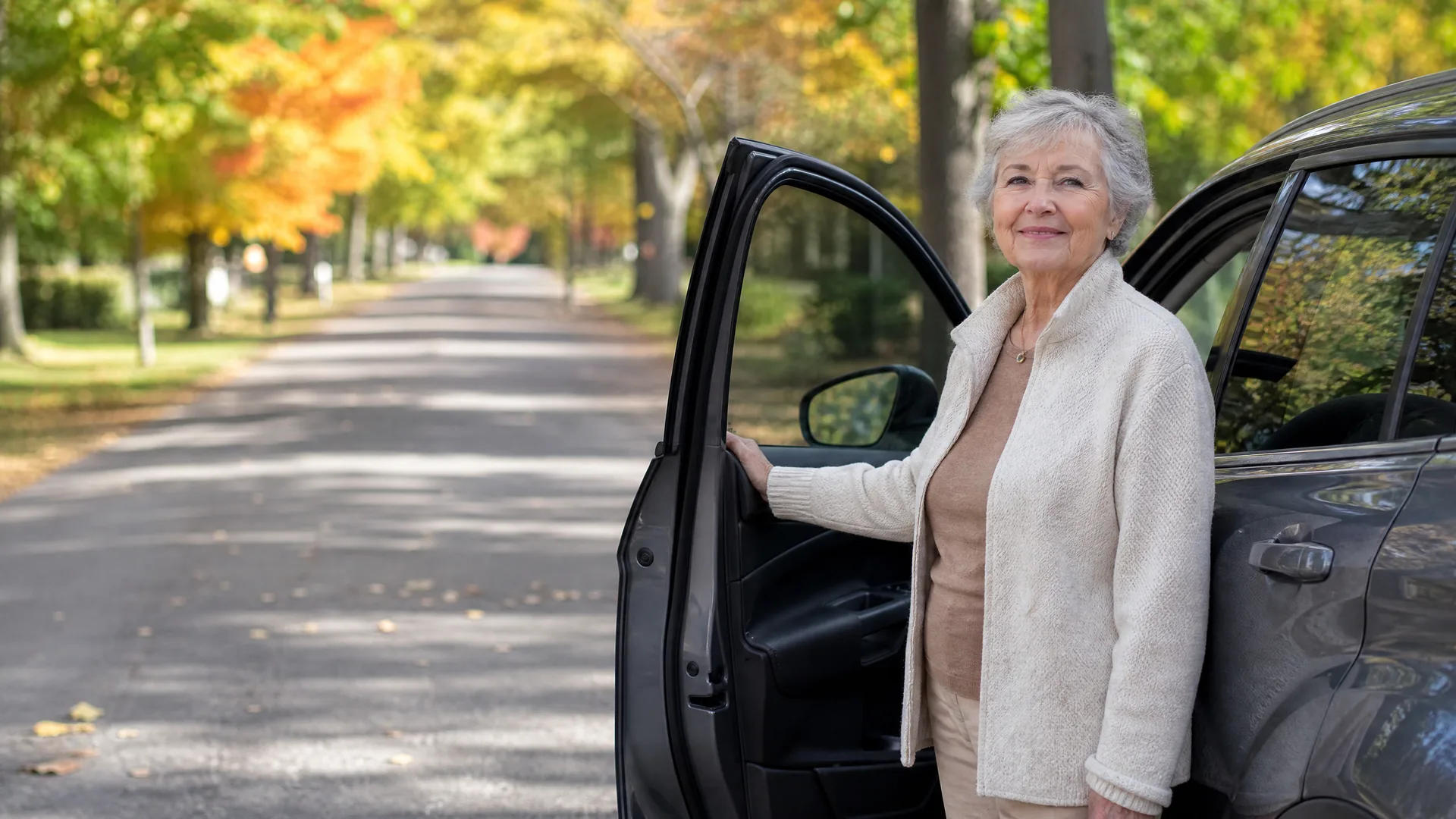 An older woman in a cream cardigan stands beside a dark car with the front passenger door open, on a tree-lined road with autumn foliage.