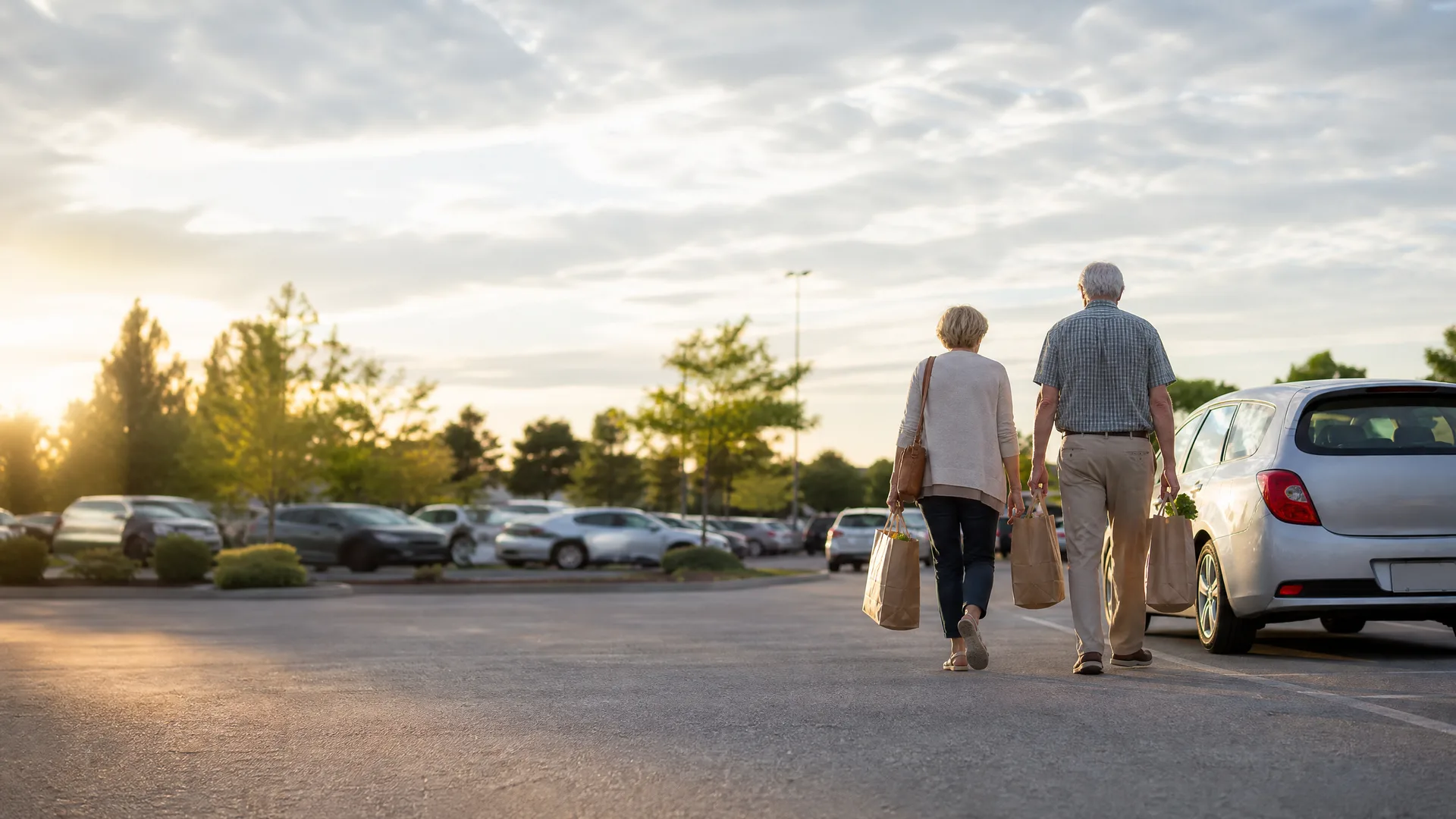 An older couple carrying paper grocery bags walks away from the camera across a parking lot at sunset, with parked cars, trees, and a silver hatchback at right.