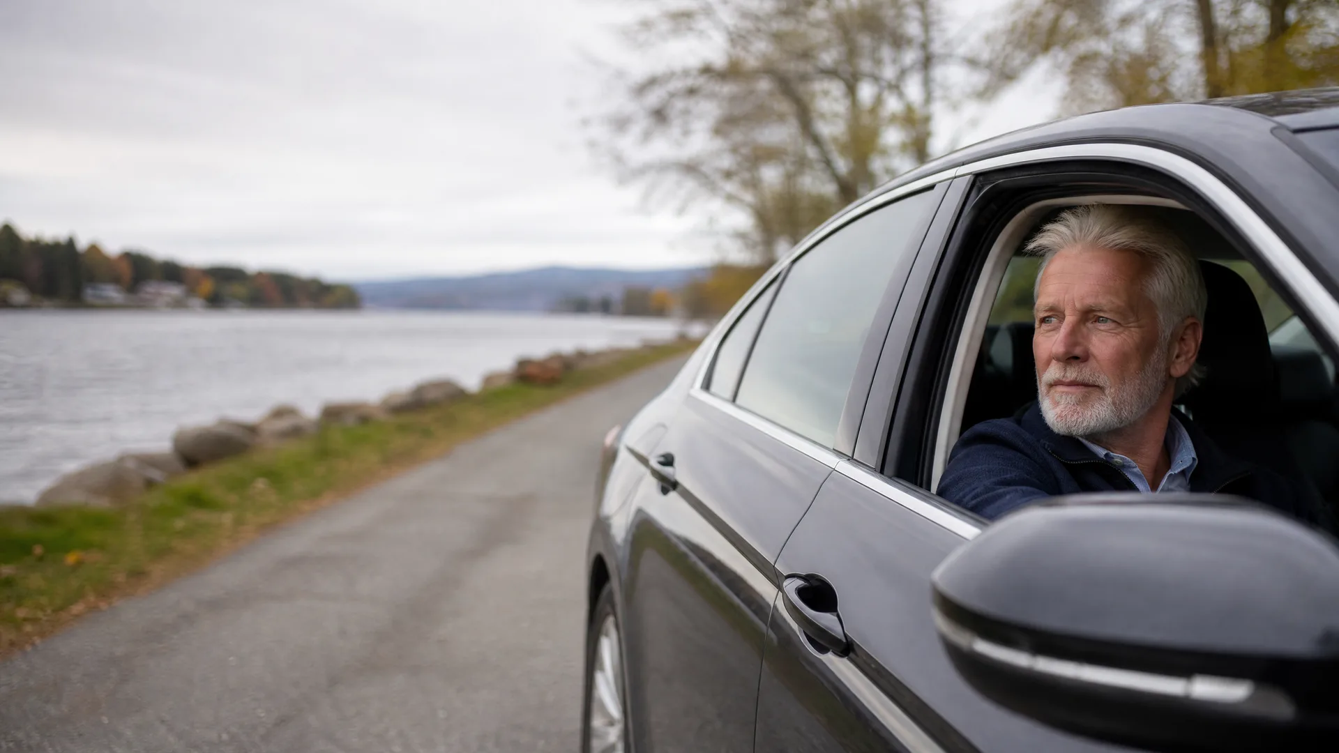 A grey-haired man with a beard looks out of the open driver's window of a dark car parked on a paved road beside a lake under an overcast sky.
