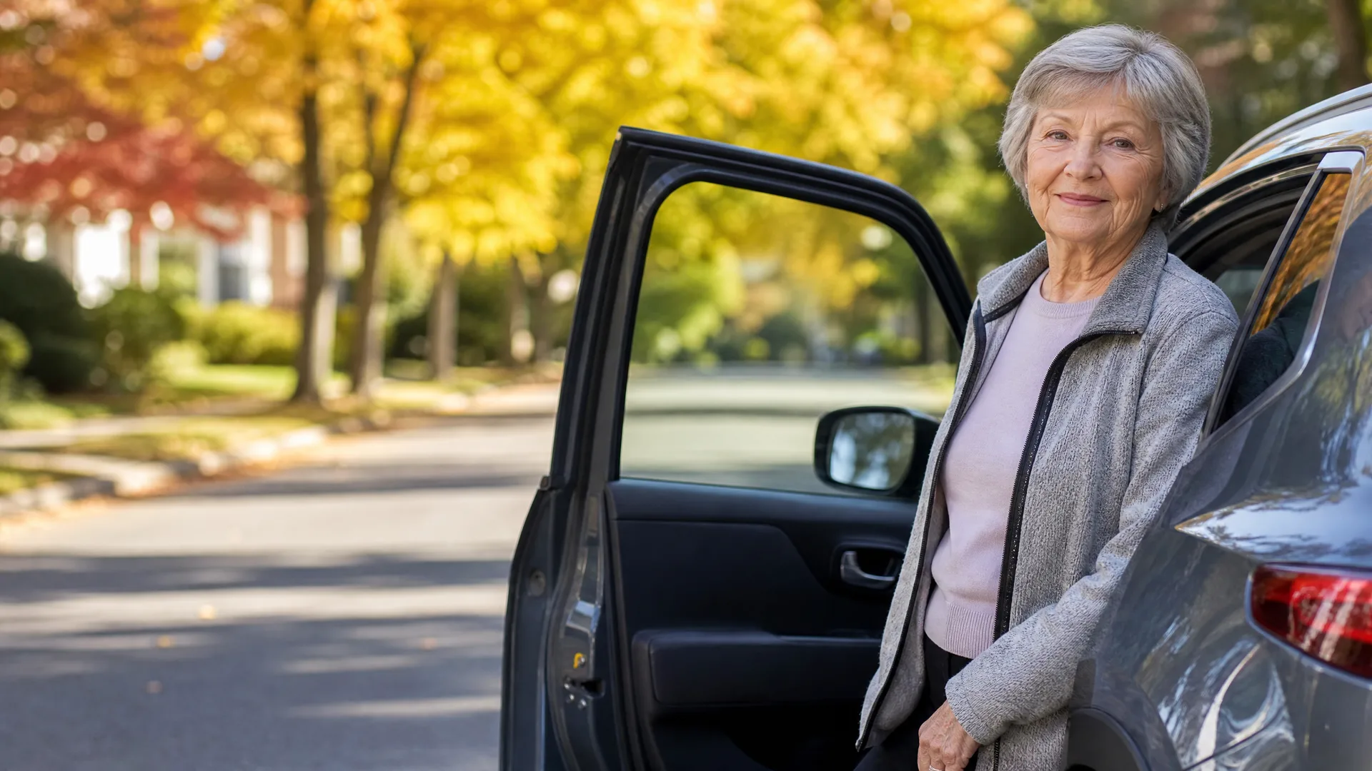 An older woman with short gray hair, wearing a gray fleece jacket over a pink top, stands beside the open driver's door of a dark car on a residential street lined with yellow and red autumn trees.