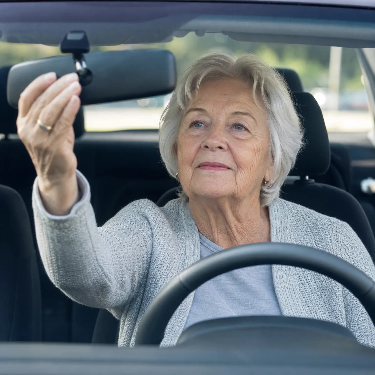An older woman with short white hair and a light gray knit cardigan sits in a car's driver seat, reaching up with her left hand to adjust the rearview mirror, with the steering wheel in the foreground and blurred greenery through the windows.