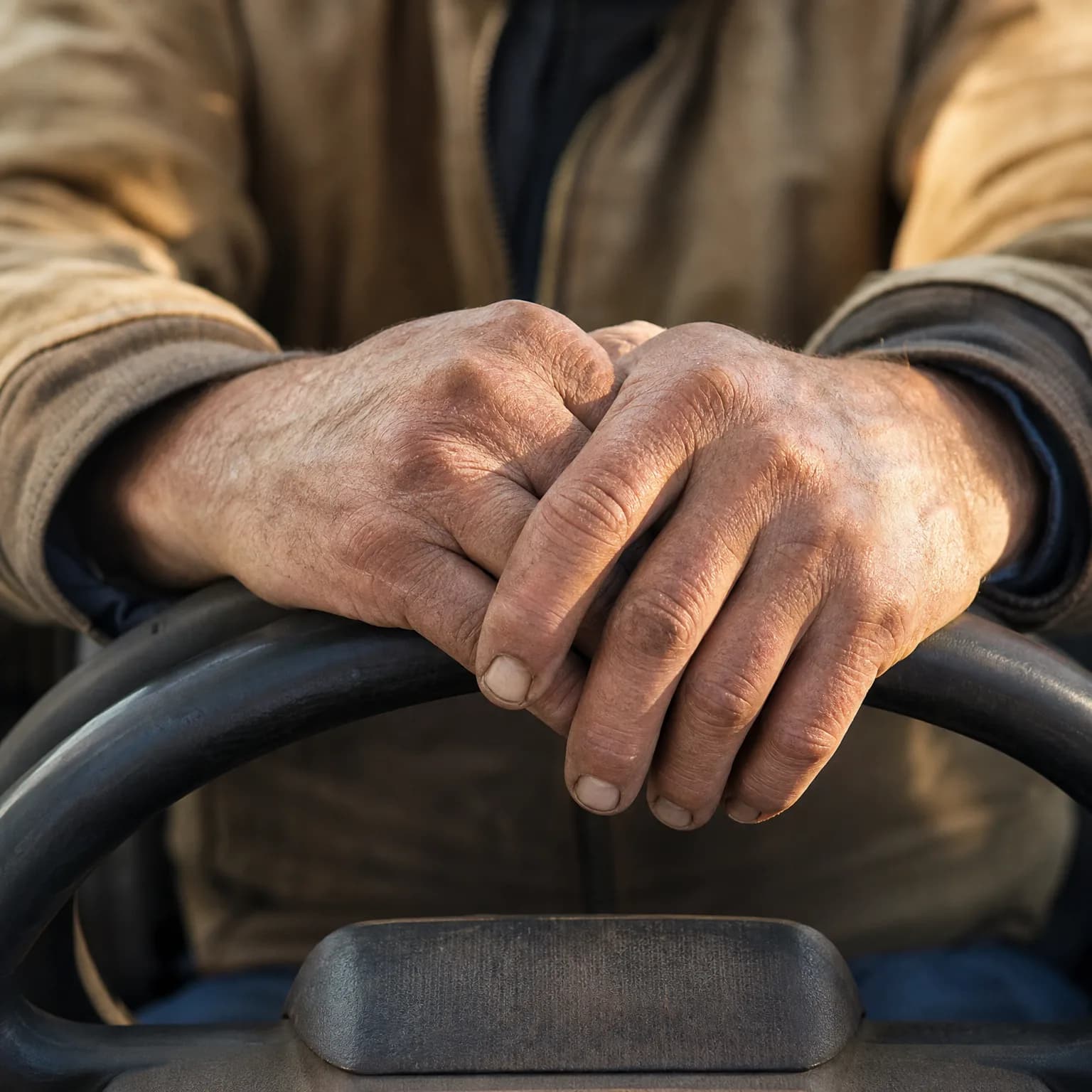 Close-up of a person's weathered, crossed hands resting on a dark steering wheel, wearing a tan jacket.