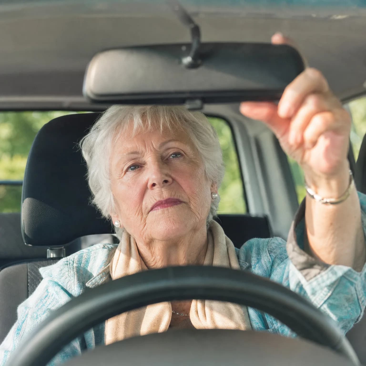 An older woman with short white hair sits in a car's driver seat, reaching up with her right hand to adjust the rear-view mirror, with the steering wheel blurred in the foreground and green foliage outside the windows.