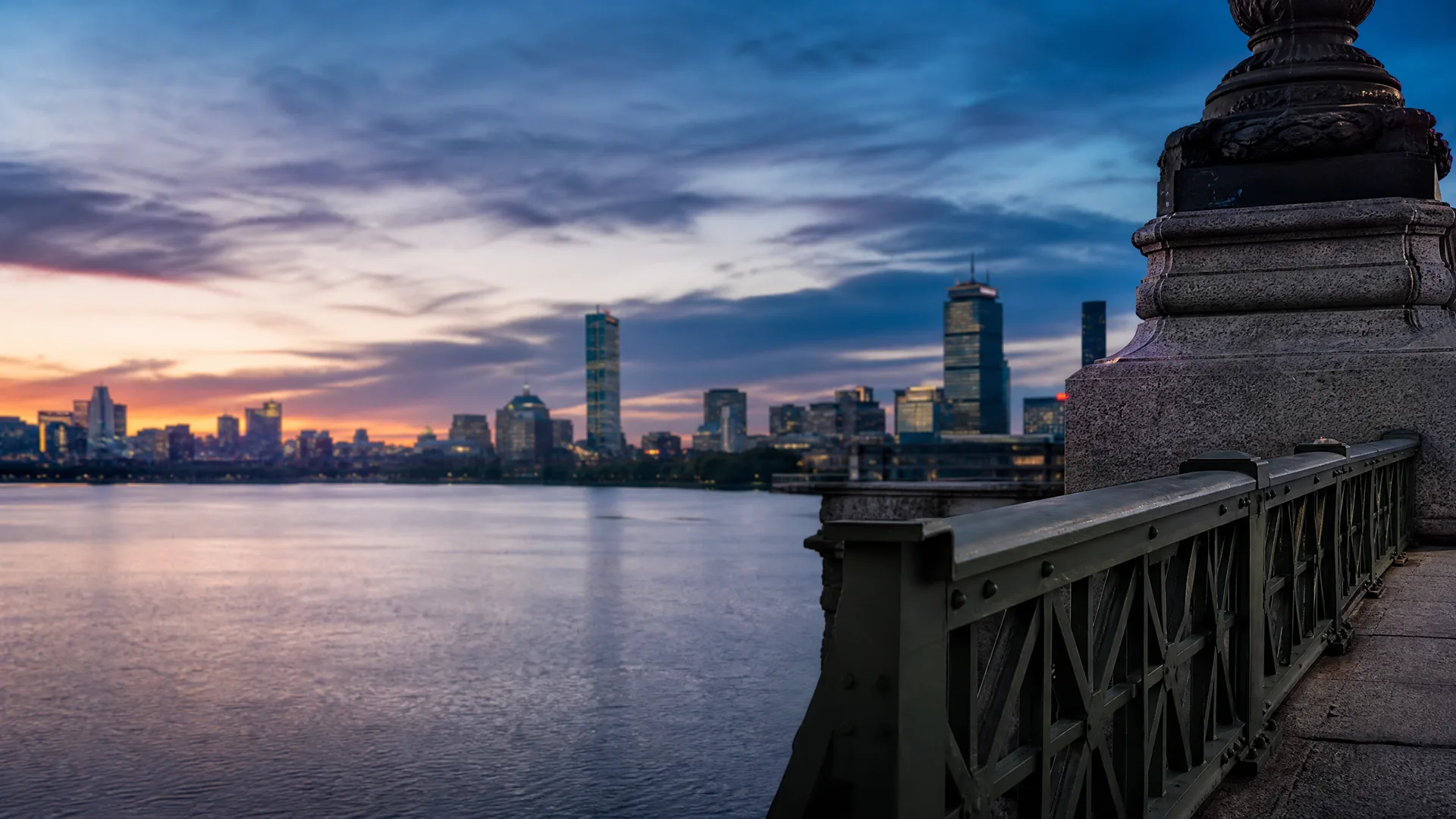 A city skyline with high-rise towers across a calm wide river at dusk, seen from a stone bridge walkway with a metal truss railing and a large stone pillar in the right foreground.
